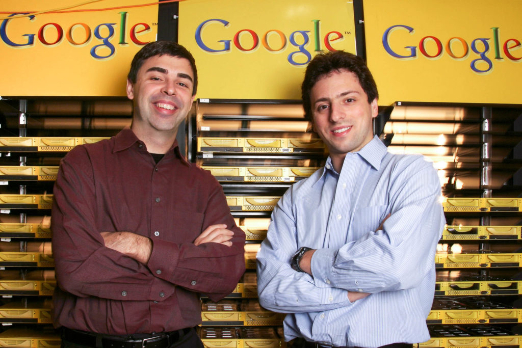 Google founders posing with smiles and folded arms in front of yellow shelves and Google signs